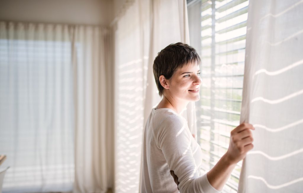 Portrait of young woman standing by window indoors at home.